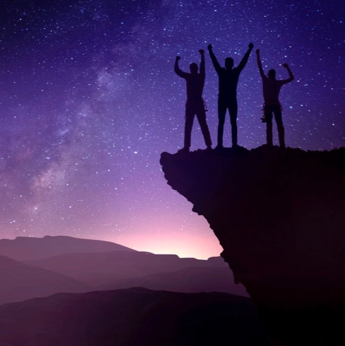 three silhouetted climbers stand arms raised after successfully working together to reach the top of a cliff